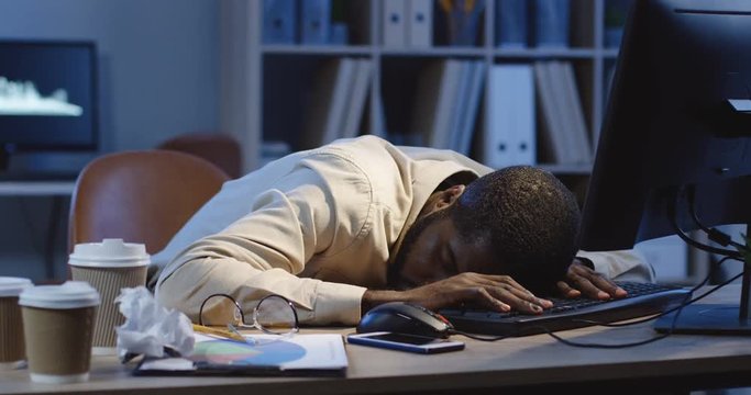African American male office worker sleeping on the table in front of the computer as being very exhausted at the night shift in the office.