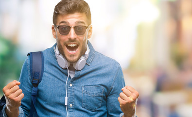 Young handsome tourist man wearing headphones and backpack over isolated background celebrating surprised and amazed for success with arms raised and open eyes. Winner concept.