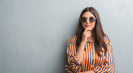 Young brunette woman over grunge grey wall wearing retro sunglasses and outfit serious face thinking about question, very confused idea