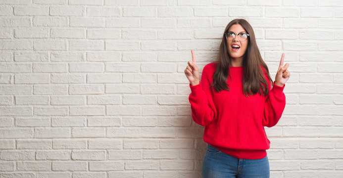 Young brunette woman standing over white brick wall amazed and surprised looking up and pointing with fingers and raised arms.