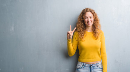 Young redhead woman over grey grunge wall showing and pointing up with fingers number two while smiling confident and happy.