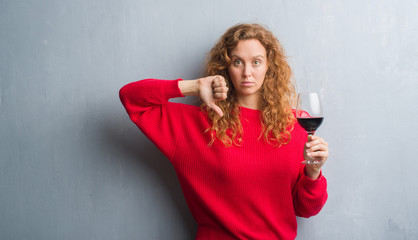 Young redhead woman over grey grunge wall drinking a glass of wine with angry face, negative sign showing dislike with thumbs down, rejection concept