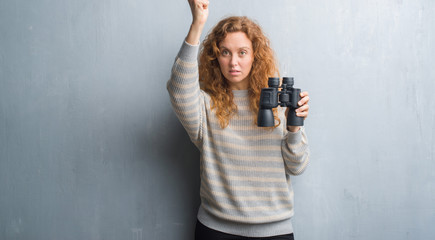 Young redhead woman over grey grunge wall looking through binoculars annoyed and frustrated shouting with anger, crazy and yelling with raised hand, anger concept