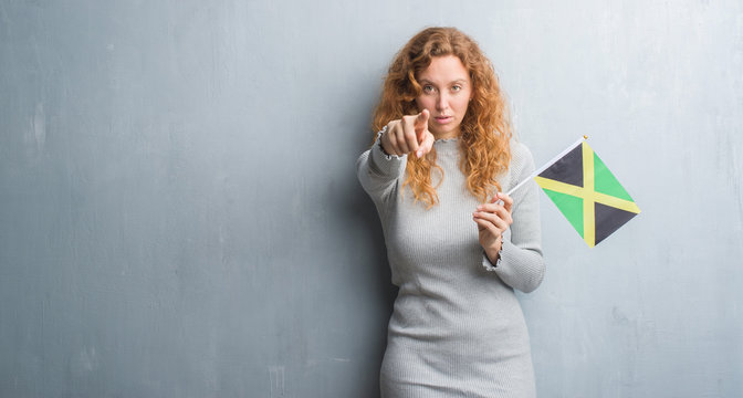 Young redhead woman over grey grunge wall holding flag of Jamaica pointing with finger to the camera and to you, hand sign, positive and confident gesture from the front