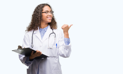 Young hispanic doctor woman holding a clipboard pointing and showing with thumb up to the side with happy face smiling