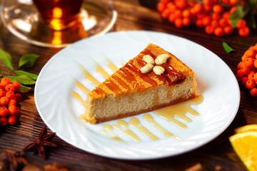 Closeup image of cheesecake slice decorated with maple syrup, cup of tea and rawan branches at wooden board background.
