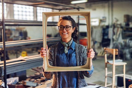 Happy creative middle age female carpenter holding picture frame in front of her face. Standing in her workshop.