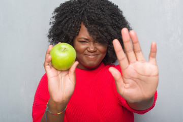 Young african american woman over grey grunge wall eating green apple with open hand doing stop sign with serious and confident expression, defense gesture