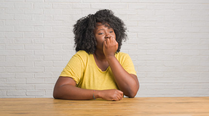 Young african american woman sitting on the table at home looking stressed and nervous with hands on mouth biting nails. Anxiety problem.