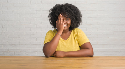 Young african american woman sitting on the table at home covering one eye with hand with confident smile on face and surprise emotion.