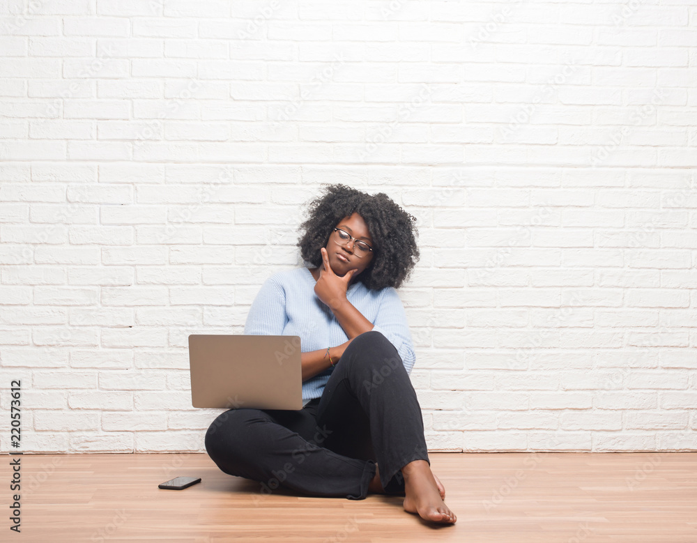 Sticker Young african american woman sitting on the floor using laptop at home with hand on chin thinking about question, pensive expression. Smiling with thoughtful face. Doubt concept.