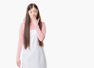 Fototapeta premium Young Chinese shop owner woman over isolated background wearing white apron cover mouth with hand shocked with shame for mistake, expression of fear, scared in silence, secret concept