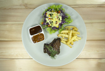 Beef steak in a white dish on a wooden background props decoration with French fries, green salad, purple cauliflower,Top view