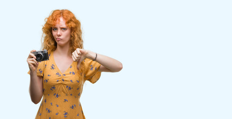 Young redhead woman taking pictures holding vintage camera with angry face, negative sign showing dislike with thumbs down, rejection concept