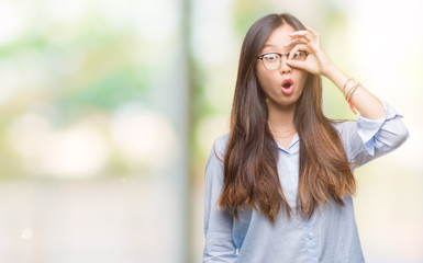 Young asian business woman wearing glasses over isolated background doing ok gesture shocked with surprised face, eye looking through fingers. Unbelieving expression.