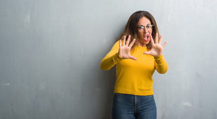 Middle age hispanic woman over grey wall wearing glasses afraid and terrified with fear expression stop gesture with hands, shouting in shock. Panic concept.