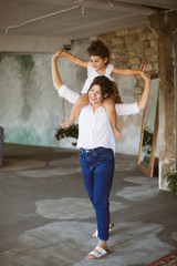 Young beautiful mother in white shirt and jeans holding little cute daughter on shoulders while dreamily spending time together at home