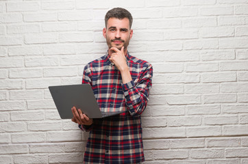 Young adult man over brick wall using laptop serious face thinking about question, very confused...