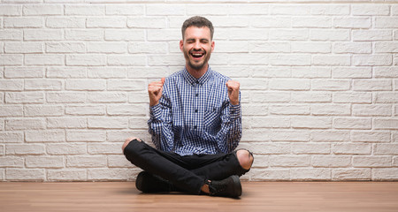 Young adult man sitting over white brick wall excited for success with arms raised celebrating victory smiling. Winner concept.