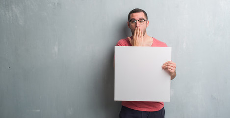 Young caucasian man over grey grunge wall holding a blank banner cover mouth with hand shocked with shame for mistake, expression of fear, scared in silence, secret concept