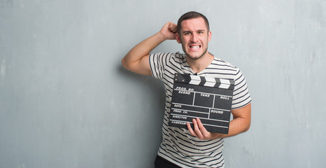 Young caucasian man over grey grunge wall holding movie clapboard annoyed and frustrated shouting with anger, crazy and yelling with raised hand, anger concept