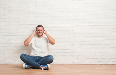 Young caucasian man sitting on the floor over white brick wall covering ears with fingers with annoyed expression for the noise of loud music. Deaf concept.