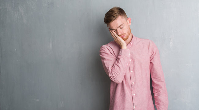 Young redhead man over grey grunge wall wearing pink shirt thinking looking tired and bored with depression problems with crossed arms.