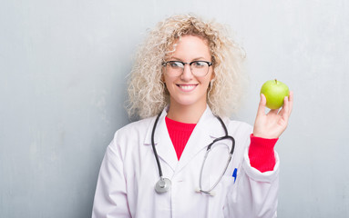 Young blonde dietitian doctor woman over grunge grey wall holding green apple with a happy face...