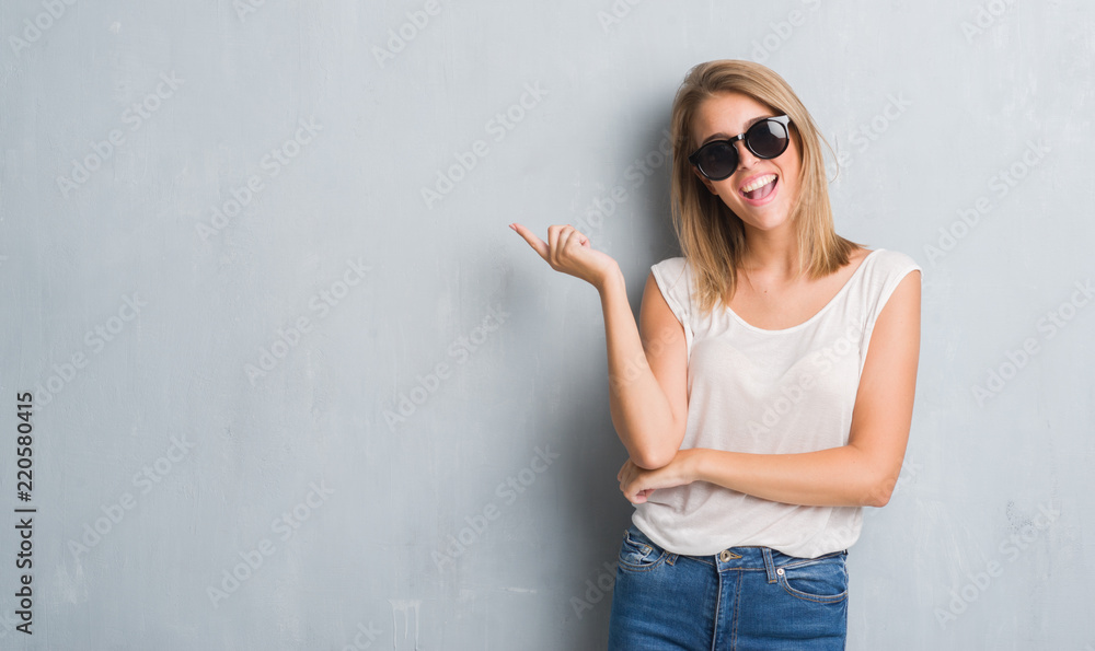 Wall mural Beautiful young woman standing over grunge grey wall wearing fashion sunglasses very happy pointing with hand and finger to the side