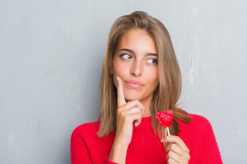 Beautiful young woman over grunge grey wall eating red heart lollipop candy serious face thinking...