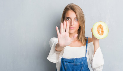 Beautiful young woman over grunge grey wall holding fresh cantaloupe melon with open hand doing...