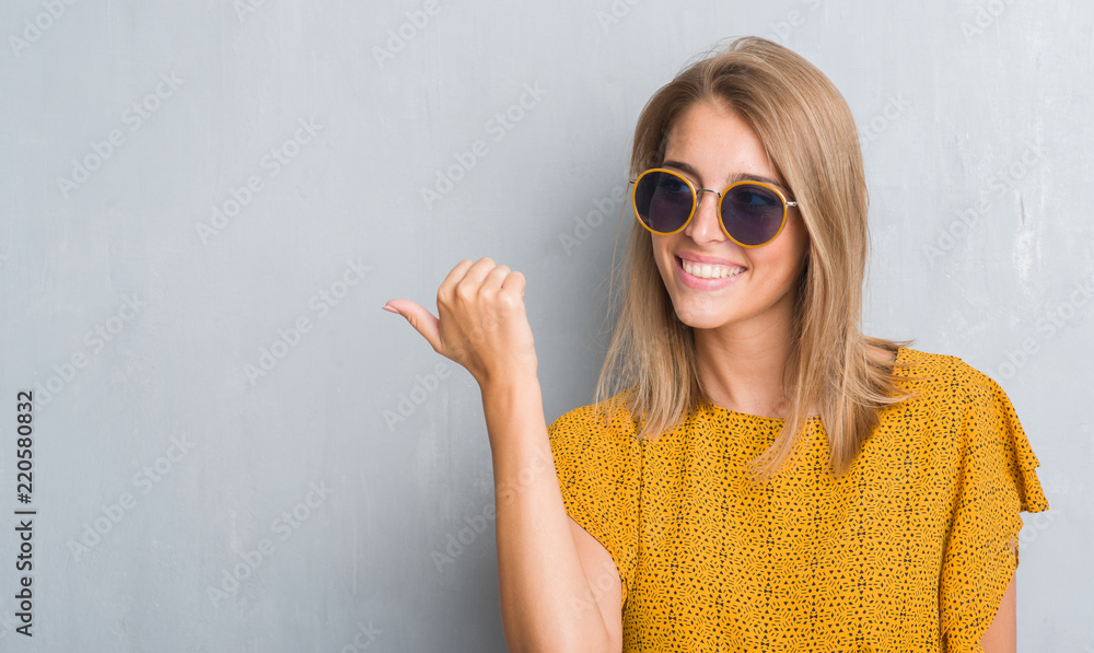 Wall mural Beautiful young woman standing over grunge grey wall wearing retro sunglasses pointing and showing with thumb up to the side with happy face smiling