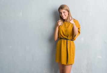 Beautiful young woman standing over grunge grey wall wearing a dress pointing fingers to camera with happy and funny face. Good energy and vibes.