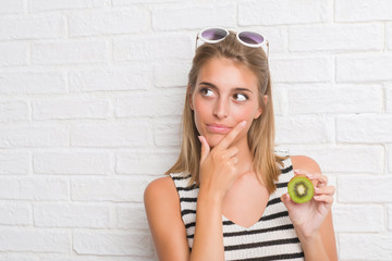 Beautiful young woman over white brick wall eating green kiwi serious face thinking about question,...