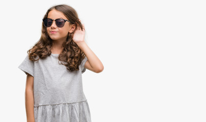Fototapeta premium Brunette hispanic girl wearing sunglasses smiling with hand over ear listening an hearing to rumor or gossip. Deafness concept.