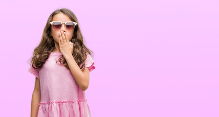 Brunette hispanic girl wearing sunglasses cover mouth with hand shocked with shame for mistake, expression of fear, scared in silence, secret concept