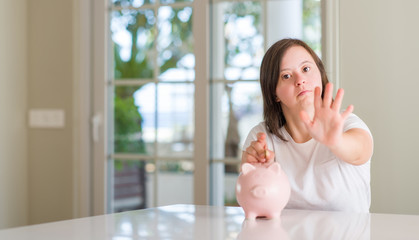 Down syndrome woman at home holding piggy bank with open hand doing stop sign with serious and confident expression, defense gesture