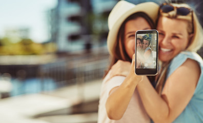 Laughing young girlfriends taking selfies together outside in su