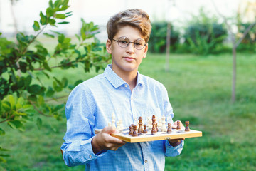 cute young boy in blue shirt holds chess board  and plays chess in the summer park. Education,...
