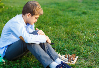 cute, young boy in blue shirt and round glasses plays on the wooden chessboard on the grass in the park. Education, hobby, intellectual game