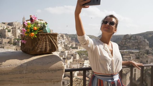 Beautiful elegant young woman in Matera Italy taking selfies pictures with smartphone sitting on a high wall viewpoint with landscape. Fashion dress with hat and skirt.