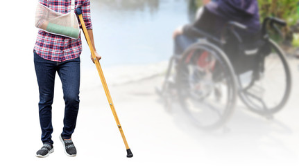 broken arm, injury woman standing wearing shirt and jeans with arm splint and holding wooden crutches isolated on blurred background patient sitting on wheelchair