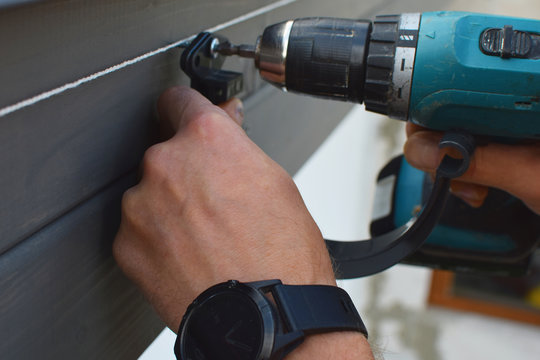 Man Hands Installing Dark Grey Plastic Roof Gutter Holder On Wooden Eaves Screwing Screws With Screwdriver For Drainage System Of Roof.