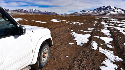 Fototapeta na wymiar A 4x4 expedition vehicle passes through the surreal winter landscapes and snow capped mountain scenery in the SIloli Desert, Sud Lipez province, Bolivia