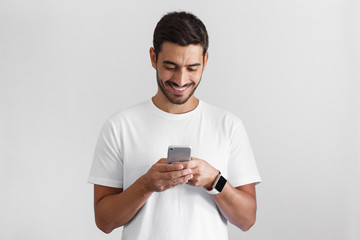 Daylight photo of young european man pictured isolated on grey background dressed in casual blank t-shirt holding smartphone with bond hands and smiling positively while looking at it
