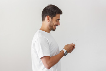 Young attractive man looking and at smartphone while texting, using mobile phone, wearing smart watches, isolated on gray background