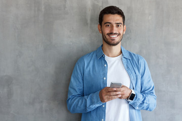 Portrait of handsome young man in blue shirt, holding smartphone, looking at camera and smiling, standing against textured wall with copy space for text