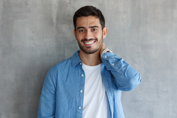 Obraz premium Indoor shot of young good looking European man standing against gray textured wall, dressed in blue shirt, showing friendly toothy smile, satisfied and confident