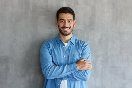 Portrait of smiling handsome man in blue shirt, standing with crossed arms against gray textured wall