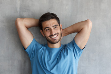 Portrait of handsome smiling young man in blue t-shirt leaning against gray textured wall with hands behind head